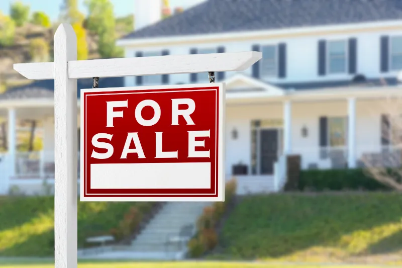 A red and white For Sale sign hangs on a post in front of a large house with a porch, windows with shutters, and a lawn. The image suggests the house is available for purchase.